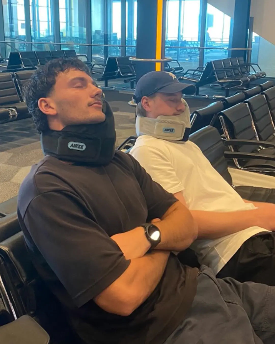Two men resting in an airport lounge using Airze travel pillows in black and grey, providing neck support and comfort for travel.